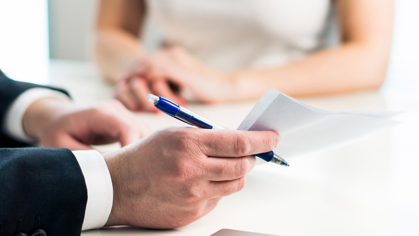 Männliche Hände mit Stift halten ein Schriftstück; ihm gegenüber sitzt eine weibliche Person, die Hände auf dem Tisch gefaltet