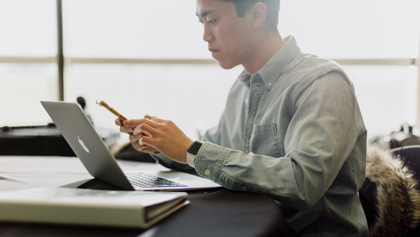 Mann sitzt grübelnd über Handy mit Laptop am Tisch